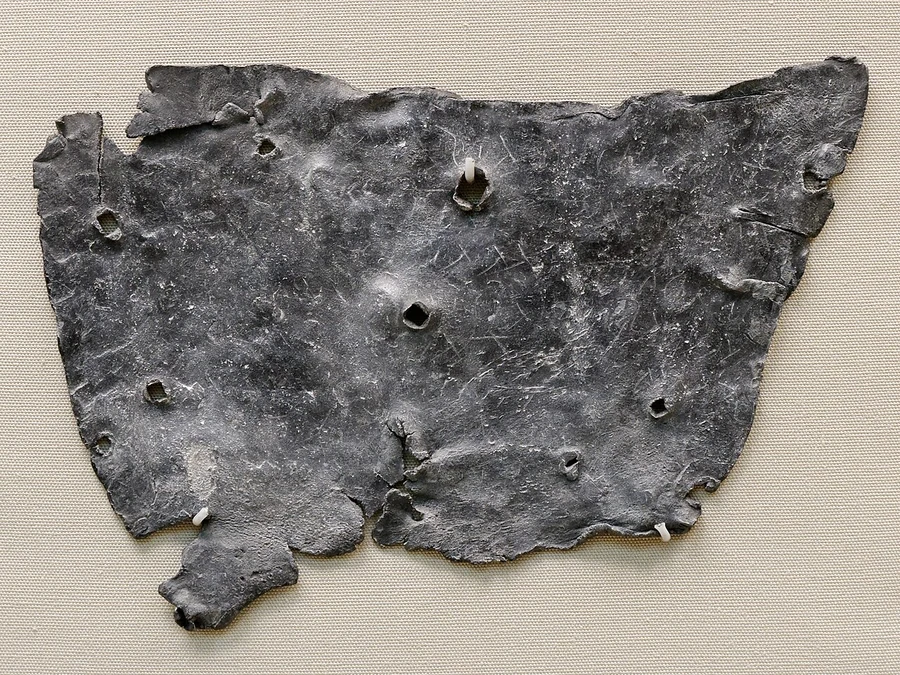 A torn, irregular sheet of corroded grey lead, mounted on linen, punctured in several places by nail holes.